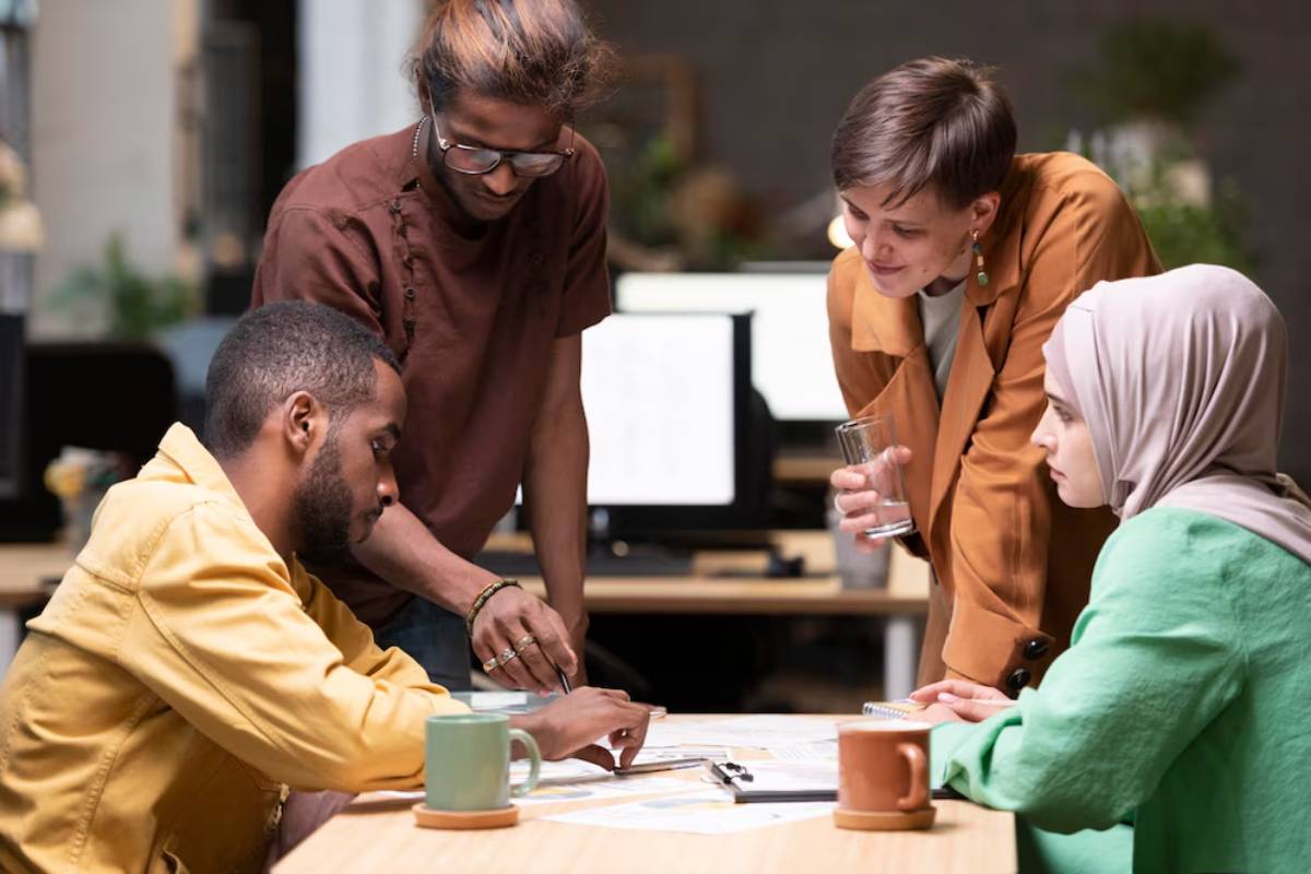 Four diverse coworkers collaborate around a table with papers and laptops in a casual office setting.