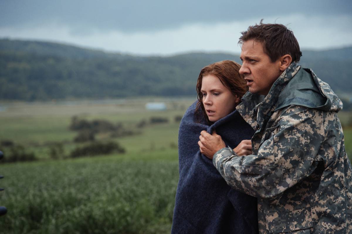 A man in a camo jacket comforting a woman wrapped in a blue blanket, both standing in a field with a cloudy backdrop.