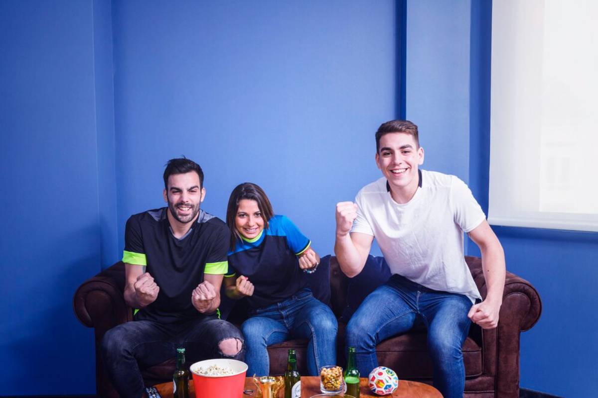 Three young adults smiling and cheering, sitting on a couch in a blue room with snacks and drinks around them.