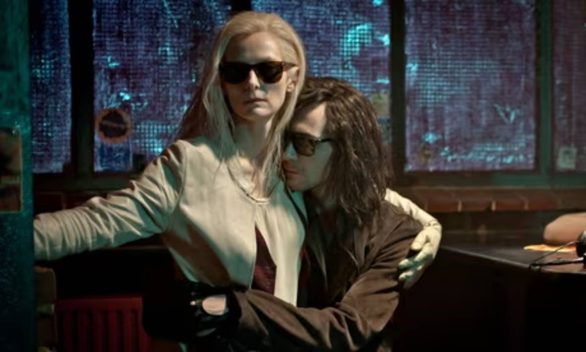 Two people in sunglasses in a dim, neon-lit room; one stands assertively while the other clings to their arm.