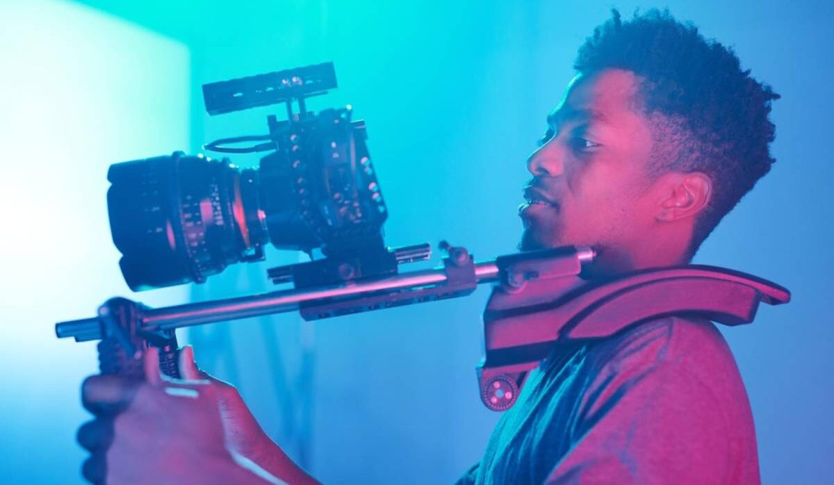 A person holds a camera rig, preparing for filming, illuminated by vibrant blue and purple lighting in a studio setting.