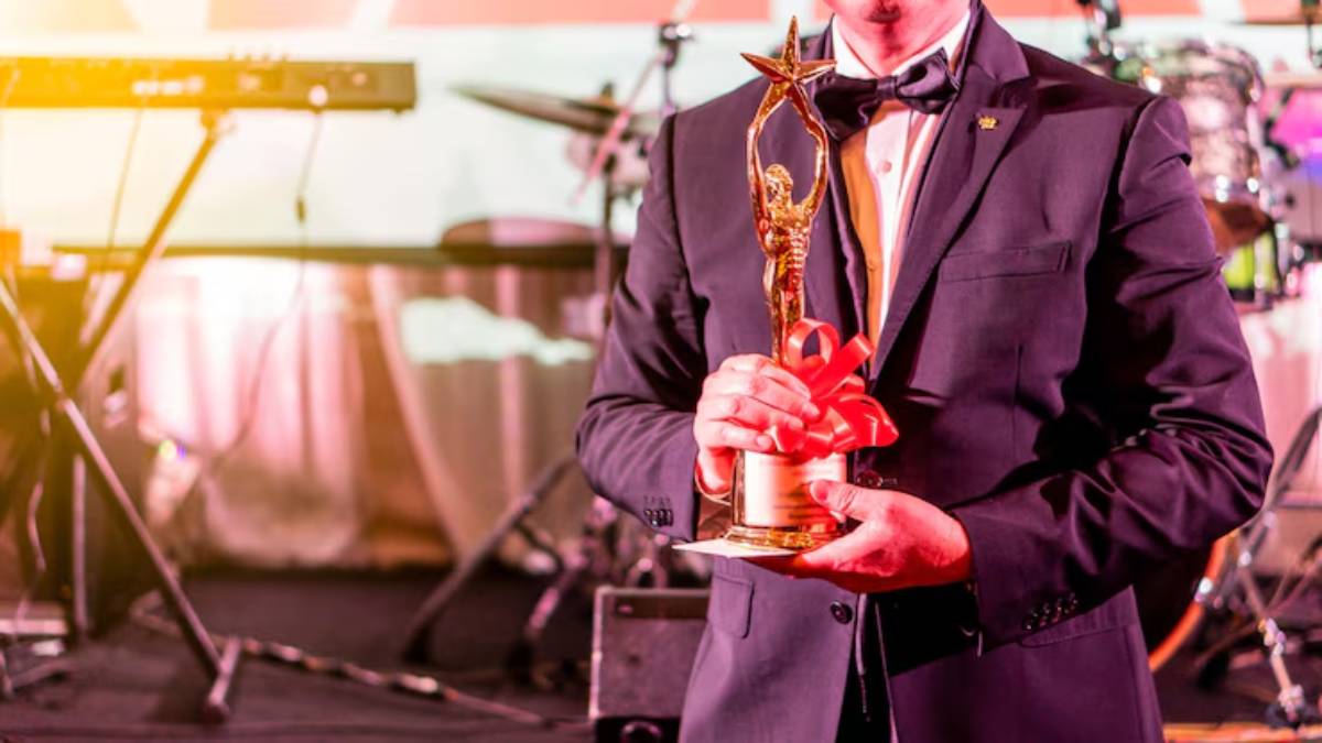 A man in a tuxedo holds a golden trophy with a star, adorned with a red ribbon, at an awards ceremony backdrop.