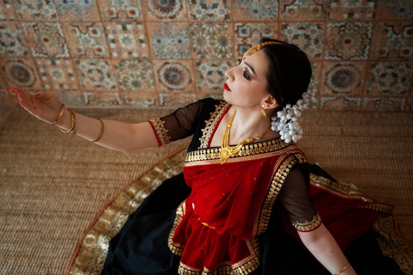 A dancer in a vibrant red and black traditional outfit gracefully poses, showcasing intricate jewelry and a floral hairpiece.