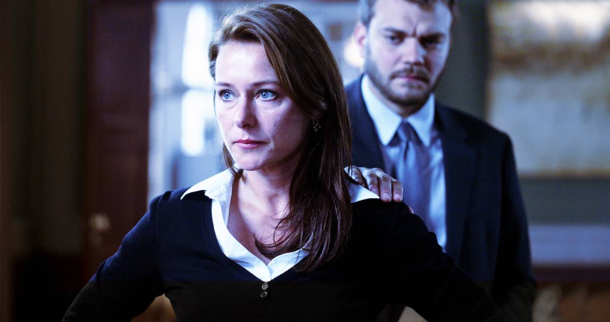A woman with long brown hair in a black blouse stands confidently, while a man in a suit has his hand on her shoulder in a dimly lit room. Scene of series - Borgen.