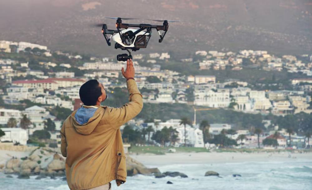 A man in a beige jacket controls a drone over a coastal city, capturing stunning views in the background.