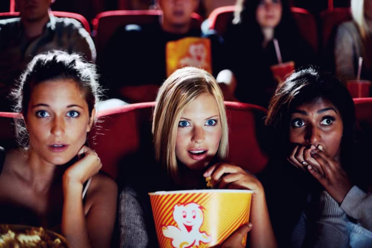 A group of moviegoers sits in a theater, enjoying popcorn and snacks, surrounded by red seats and dim lighting.