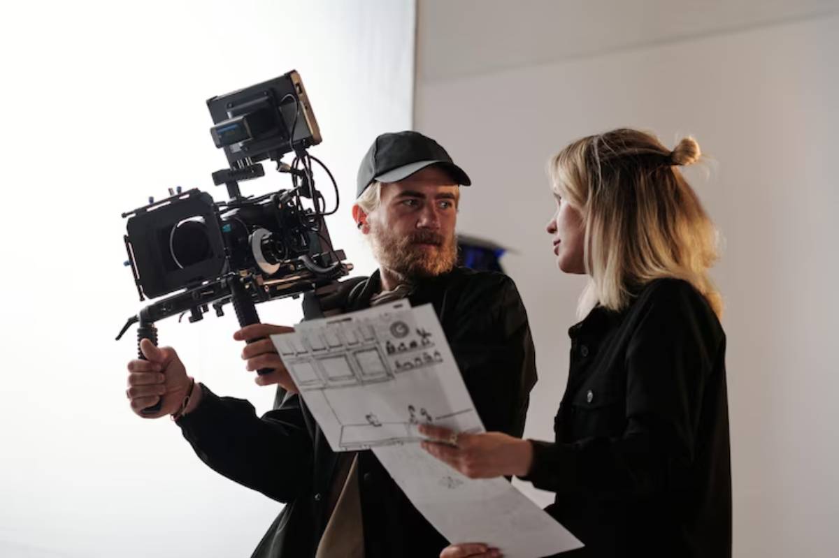 A film crew member holds a camera rig while reviewing a storyboard with director in a well-lit studio setting.