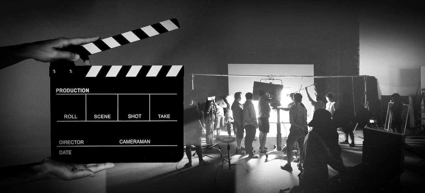A film clapperboard held in the foreground with a busy movie set in the background, featuring crew members preparing for a shoot.