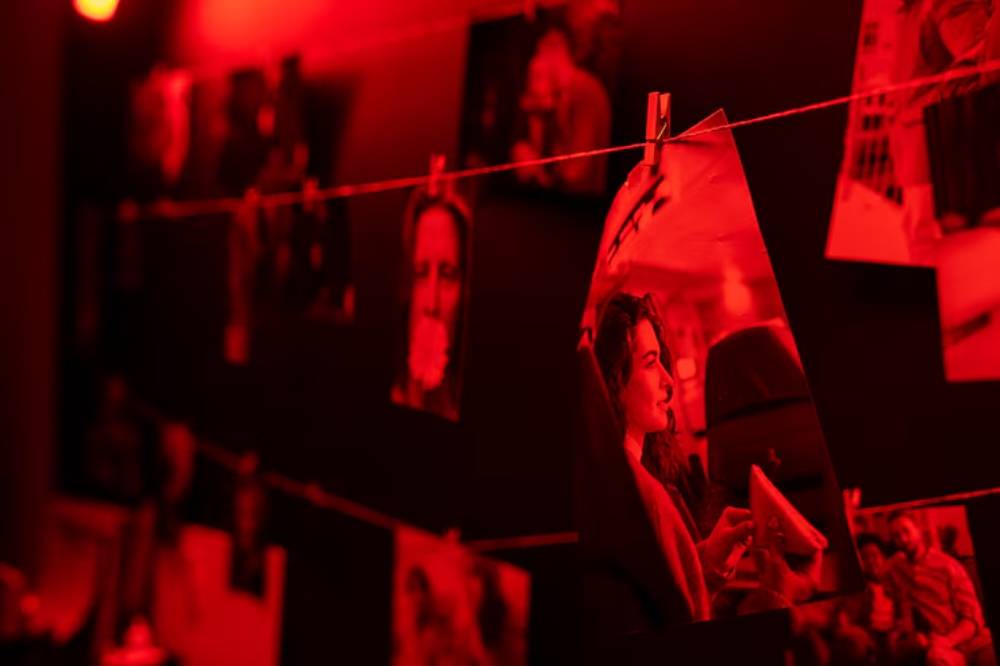 A collection of black and white photographs hangs from a string, illuminated by a dramatic red light, creating a moody atmosphere.