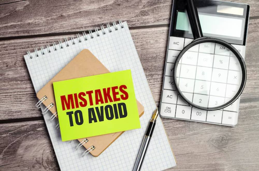 A bright yellow sticky note with 'MISTAKES TO AVOID' sits on a notebook and next to a calculator and magnifying glass on a wooden table.
