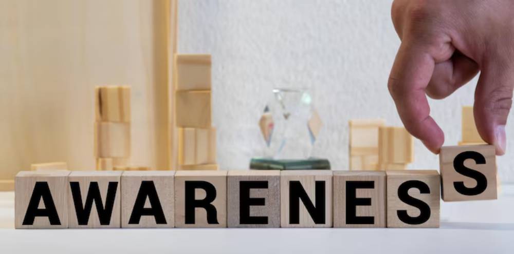 A hand positions a wooden block to complete the word 'AWARENESS'. set against a backdrop of other wooden blocks and decorative objects.