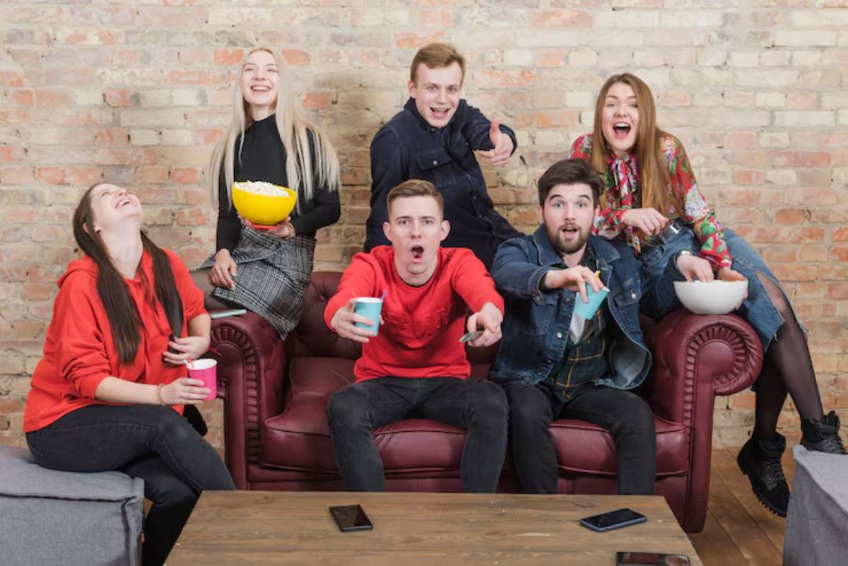 A group of six friends gathers on a burgundy couch and watching movie, enjoying snacks and drinks in a cozy, rustic room with a brick wall.