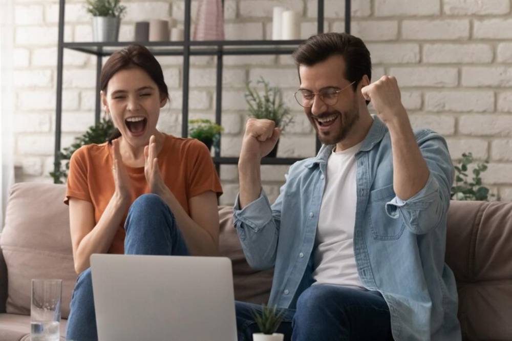 A man and woman excitedly celebrate together on a couch, with a laptop in front of them and a cozy, modern background.