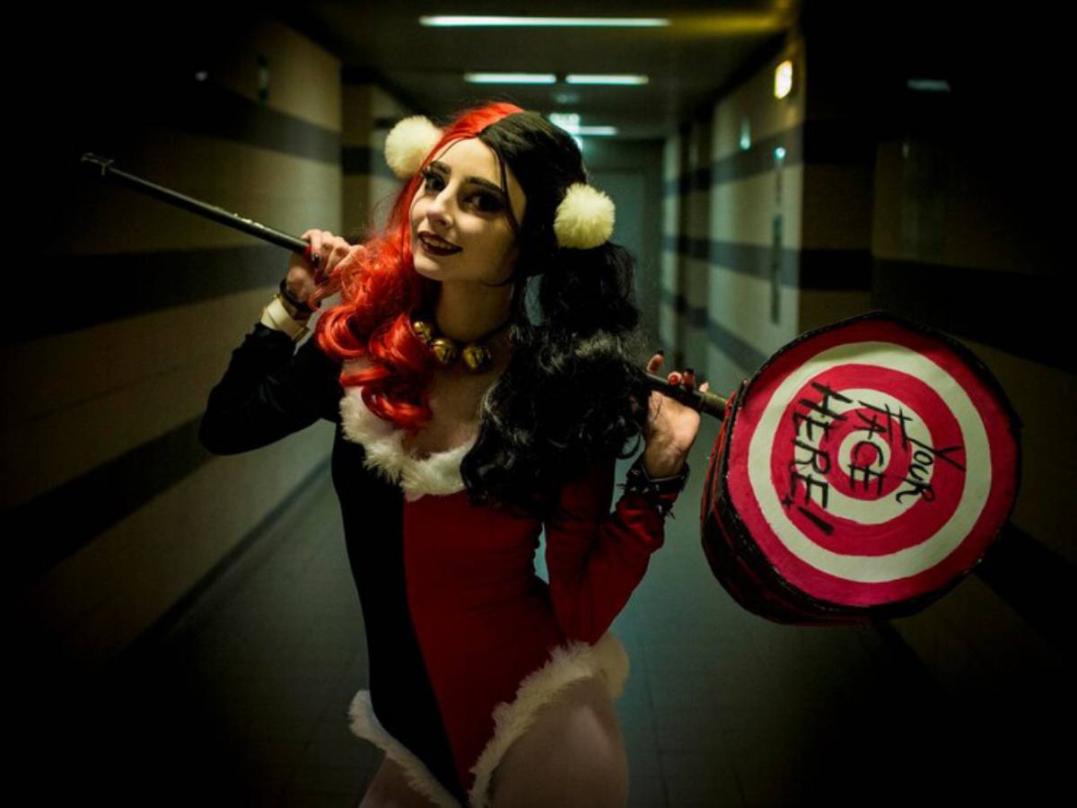 A woman in a vibrant red and black costume holds a large, decorative mallet with a bullseye design, standing in a dimly lit hallway.