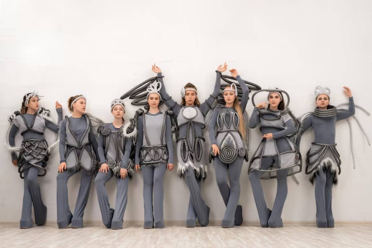 A group of eight people dressed in unique gray outfits with intricate designs and textures, striking a dynamic pose against a white wall.