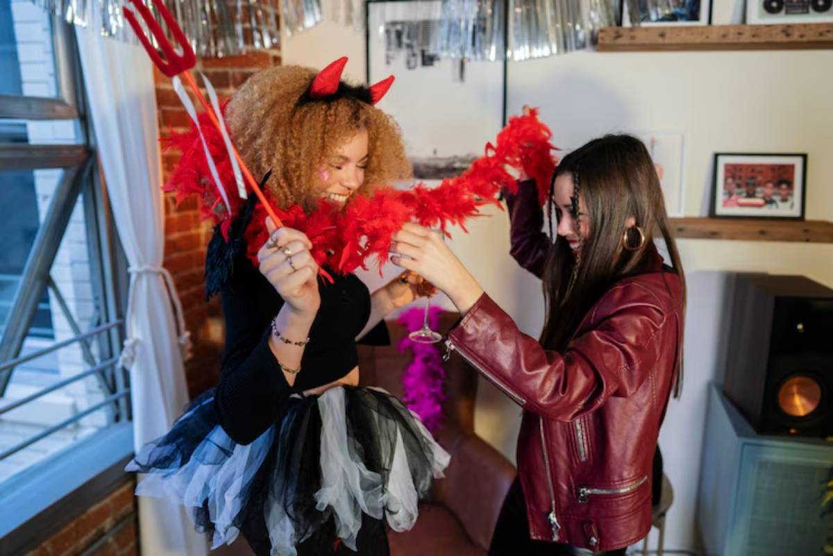 Two women celebrate in a colorful room, one dressed as a devil with horns and a red feather boa, enjoying drinks and festive decor.
