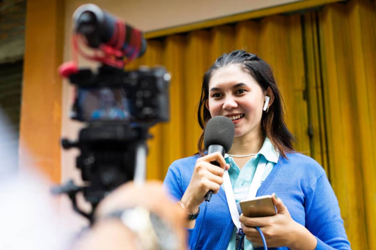 A woman holding a microphone and smartphone, wearing earbuds, stands in front of a yellow backdrop while being filmed in a film festival.
