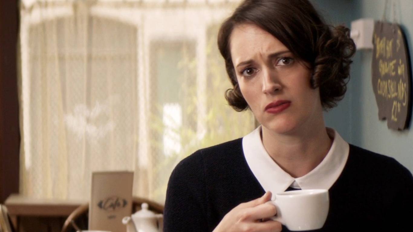A person in a black sweater with a white collar holds a cup of coffee, seated in a cozy cafe with soft daylight filtering through curtains. Scene of series - Fleabag .