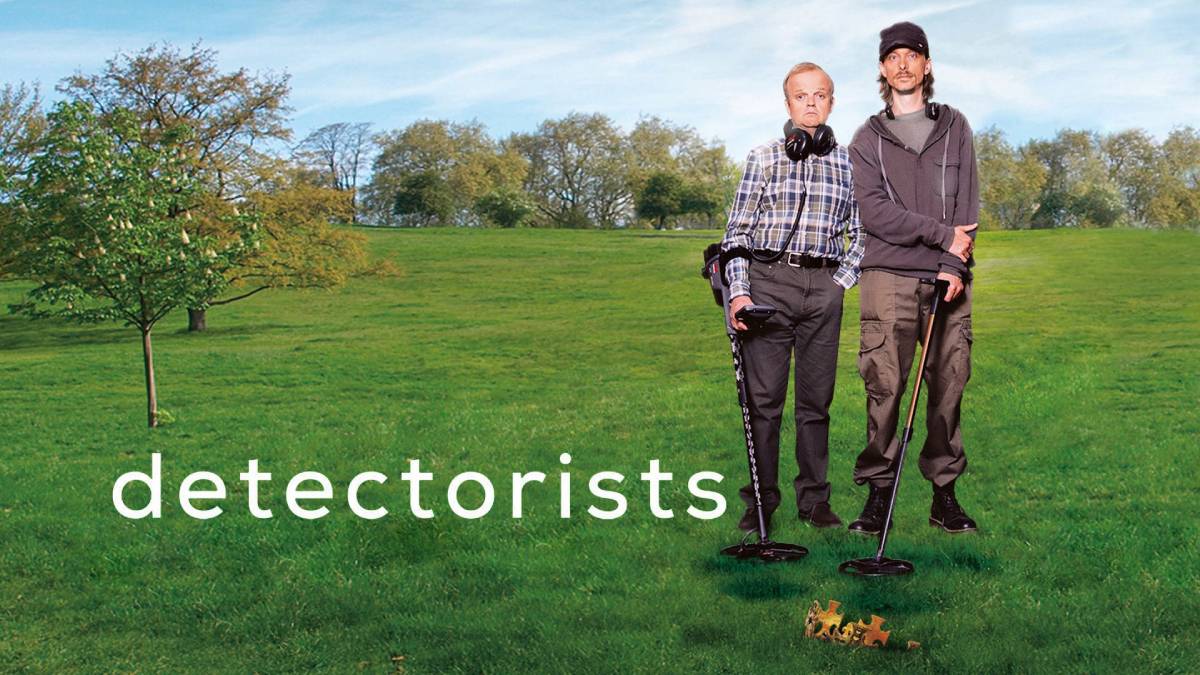 Two individuals stand on a lush green field, equipped with metal detectors, ready for a treasure hunt.
