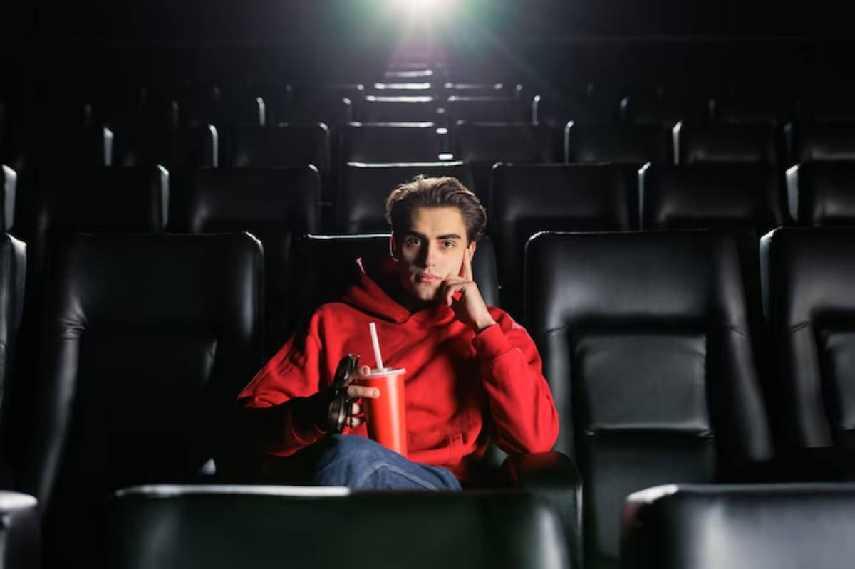 A person in a red hoodie sits in a movie theater and watching movie, holding a drink, surrounded by empty black seats and a bright light in the background.