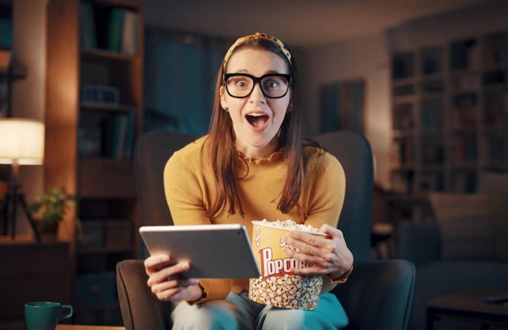 A person in a cozy living room watching movie and sits on a chair, holding a tablet in one hand and a bucket of popcorn in the other.