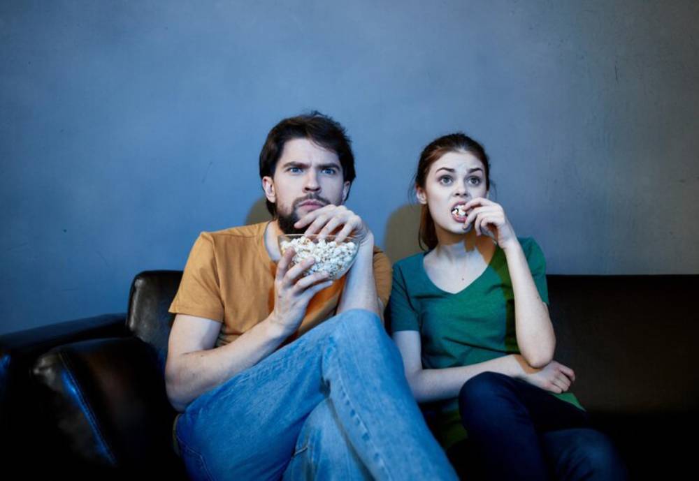 A person in an orange shirt holds a bowl of popcorn, sitting beside another in a green shirt on a dark sofa against a gray wall and watching series together