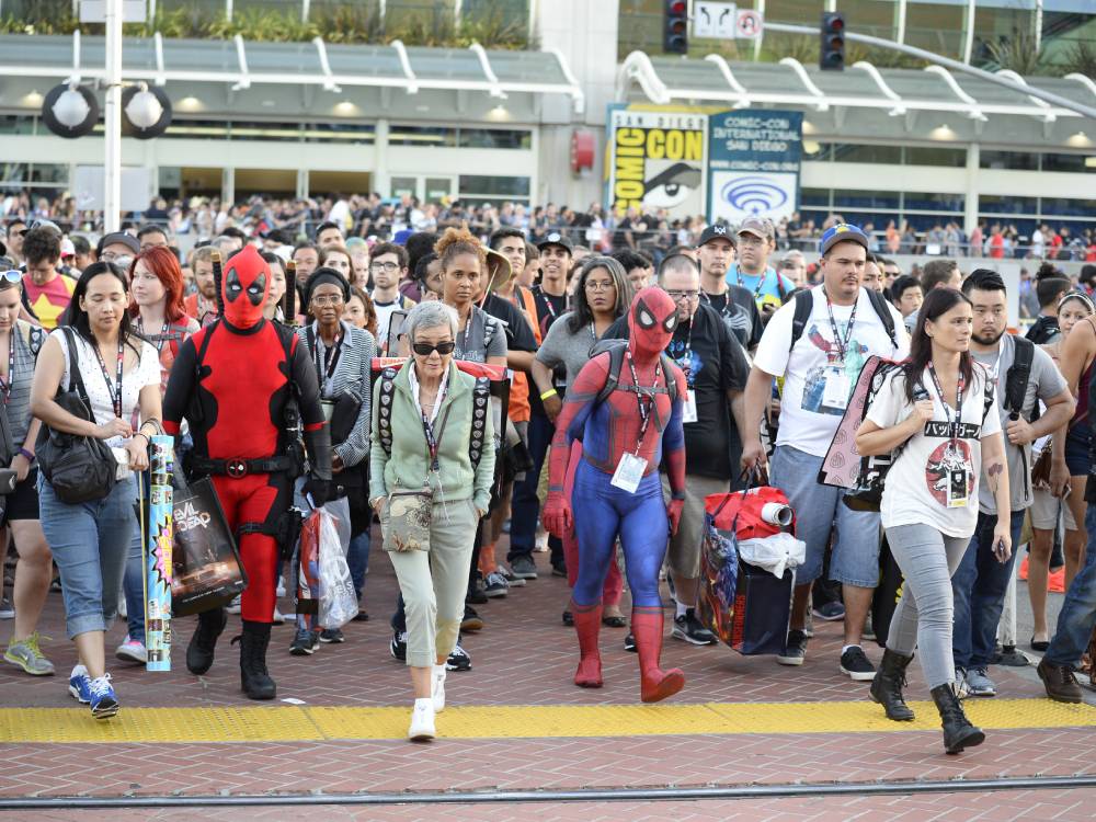 A crowd of comic book fans and cosplayers, including Deadpool and Spider-Man, walk towards the convention center at Comic-Con.