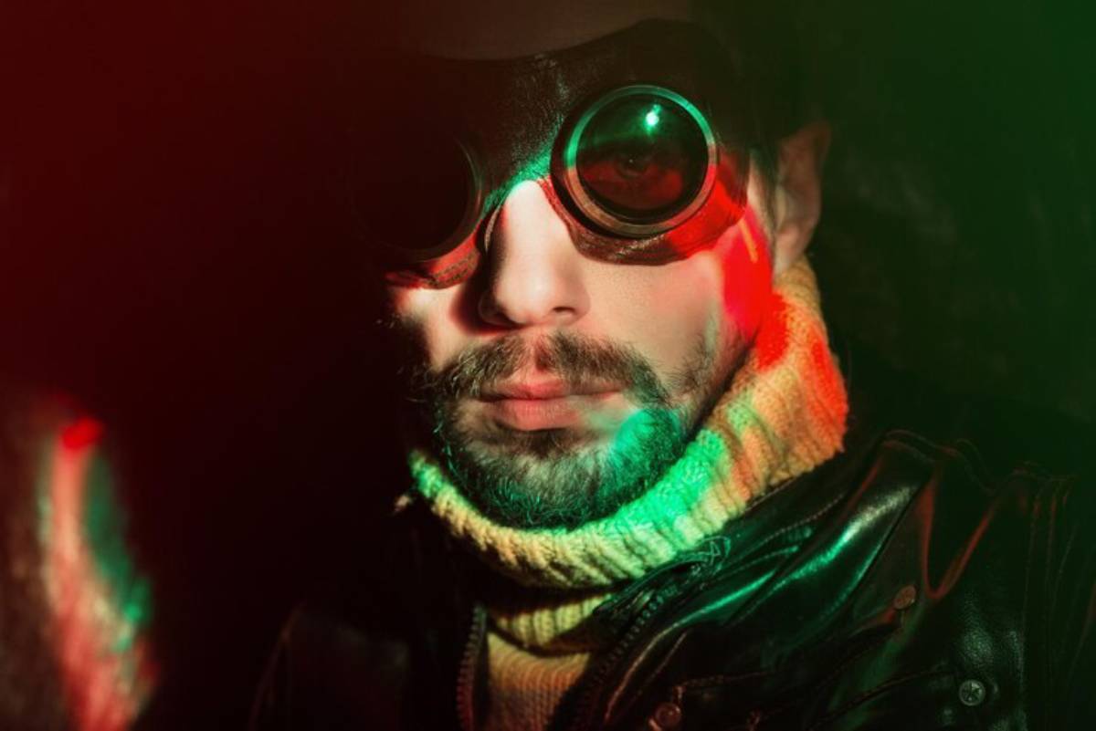 Cinematic shot of a person in a black leather jacket and colorful sweater stands against a dark background with vibrant green and red lighting.