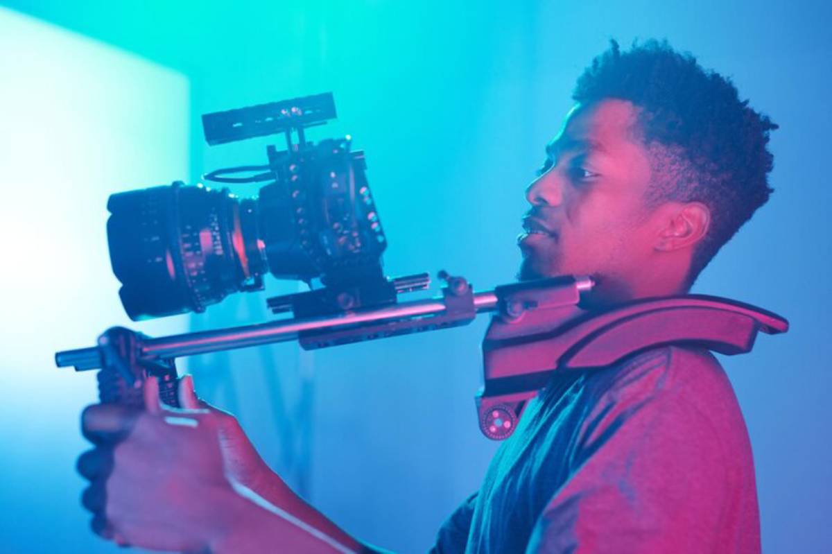 A person holds a professional camera rig with a large lens, surrounded by soft blue and purple lighting in a studio setting.