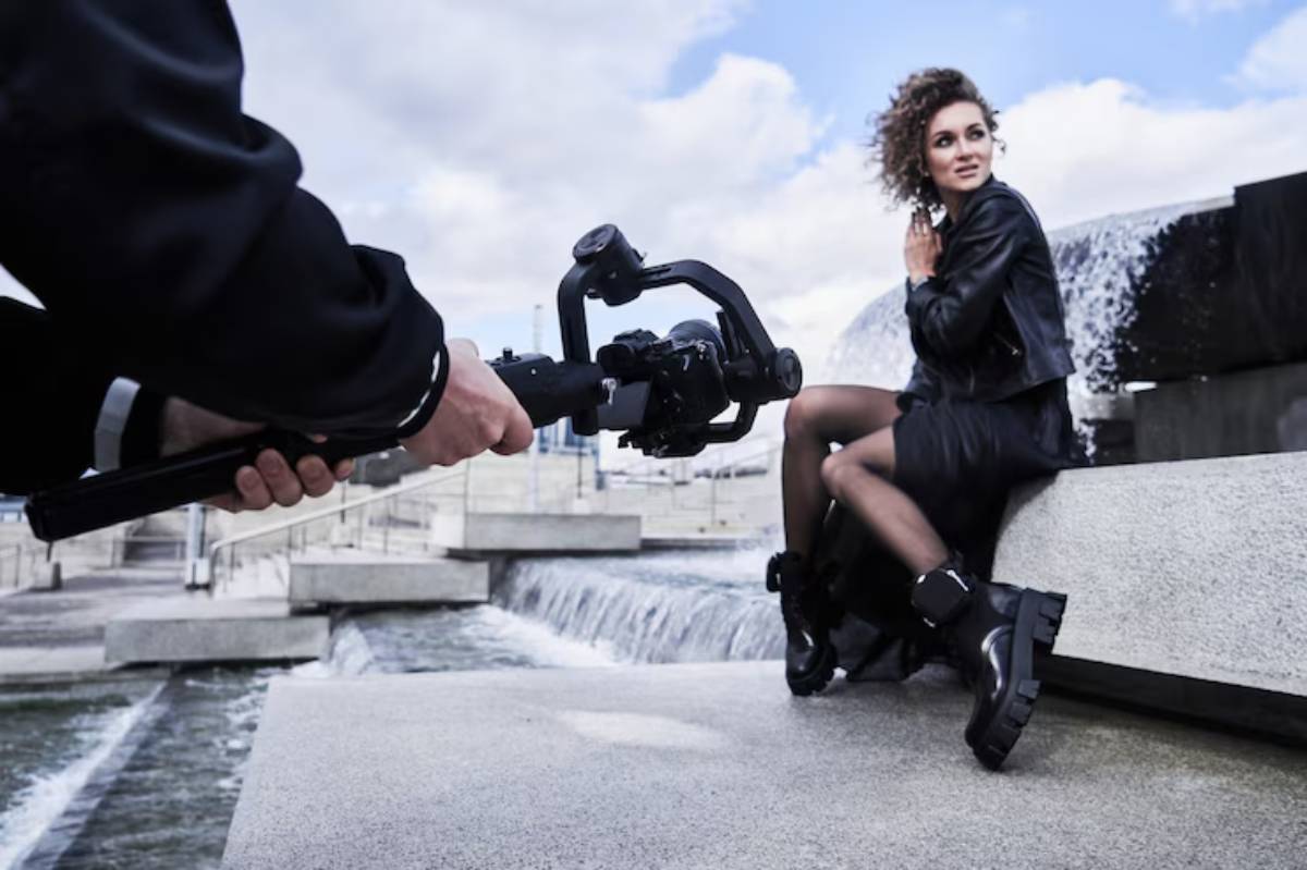 A person captures a model posing near a waterfall, showcasing stylish attire and footwear, with clouds rolling in the background.