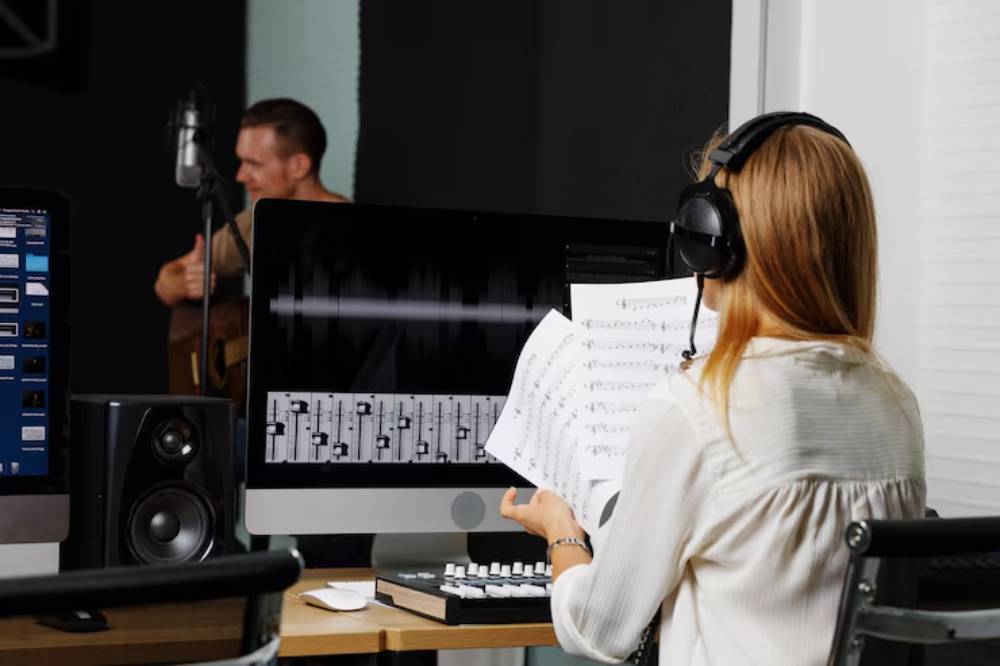 A woman wearing headphones reviews sheet music in a music studio, with computers and audio equipment visible in the background.