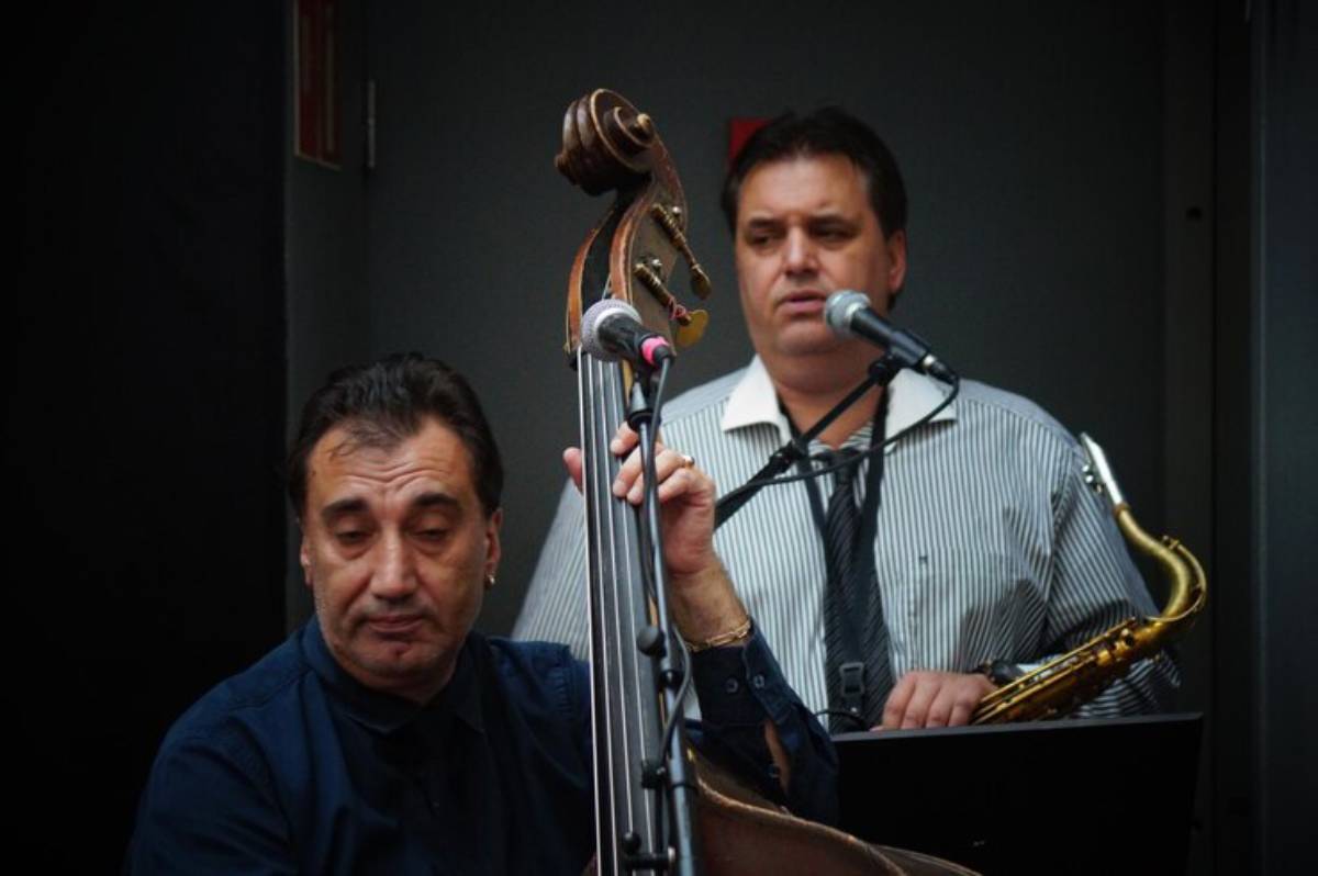 A musician plays a double bass while another stands behind a microphone holding a saxophone, creating a lively musical atmosphere.