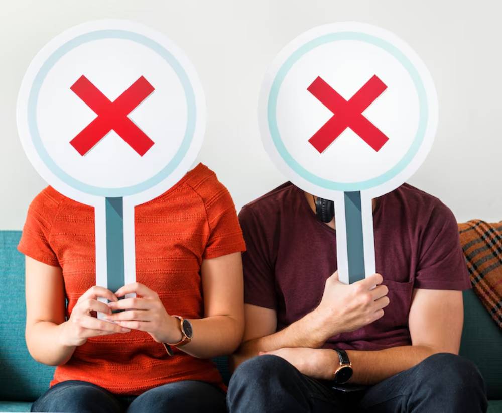 A man and woman hold large signs with red Xs, obscuring their faces, while sitting on a couch against a neutral background.