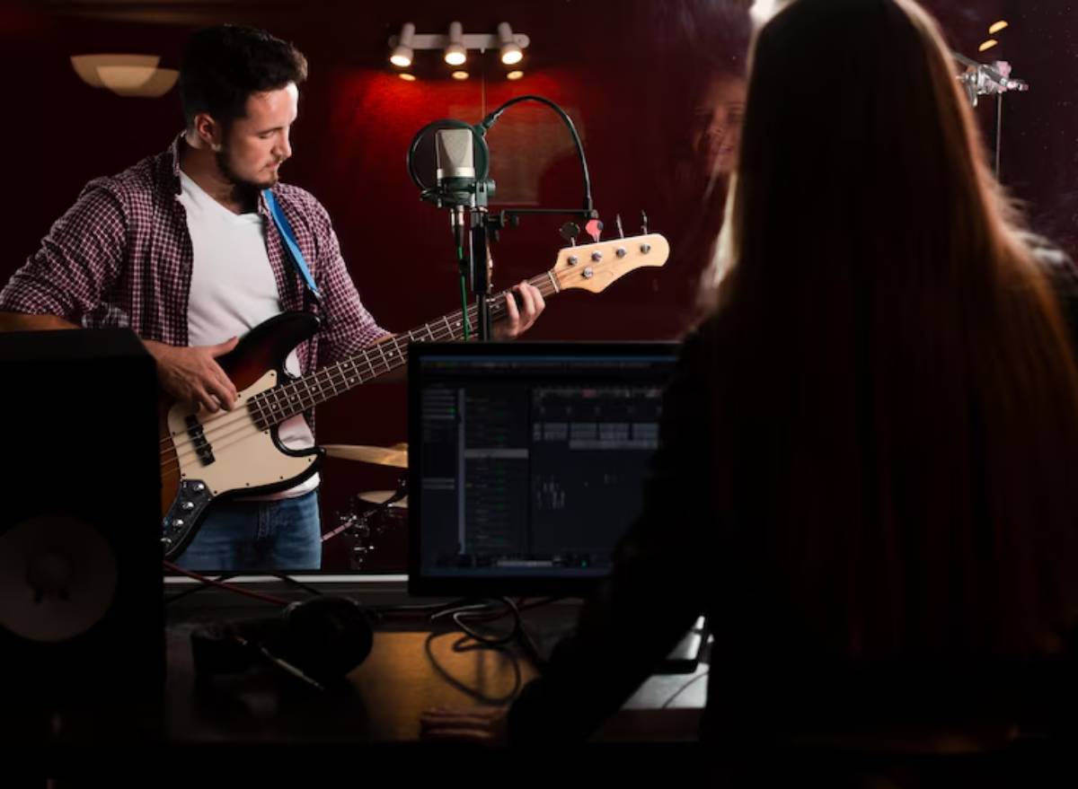 A musician plays bass guitar in a recording studio, while a producer focuses on music software at a computer.